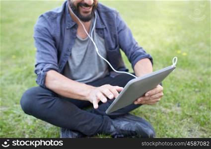 people, music, technology, leisure and lifestyle - close up of happy hipster man with tablet pc and earphones sitting on grass