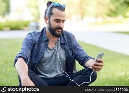 people, music, technology, leisure and lifestyle - happy young hipster man with earphones and smartphone sitting on grass