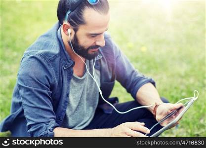 people, music, technology, leisure and lifestyle - happy young hipster man with earphones and tablet pc computer sitting on grass. man with earphones and tablet pc sitting on grass. man with earphones and tablet pc sitting on grass