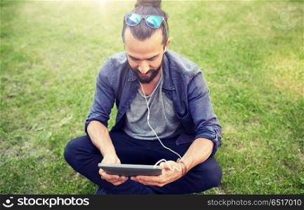 people, music, technology, leisure and lifestyle - happy young hipster man with earphones and smartphone sitting on grass. man with earphones and smartphone sitting on grass. man with earphones and smartphone sitting on grass