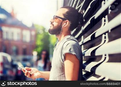 people, music, technology, leisure and lifestyle - happy young hipster man with earphones and smartphone on city street. happy man with earphones and smartphone in city. happy man with earphones and smartphone in city