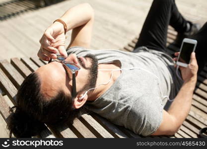 people, music, technology, leisure and lifestyle - happy young hipster with earphones and smartphone lying on city street bench