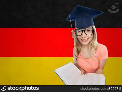 people, national education, knowledge and graduation concept - smiling young student woman in mortarboard and eyeglasses with encyclopedia book over german flag background