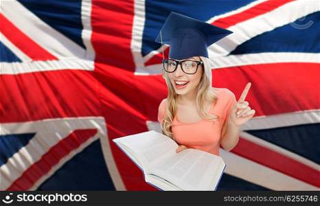 people, national education, knowledge and graduation concept - smiling young student woman in mortarboard and eyeglasses with encyclopedia book pointing finger up over english flag