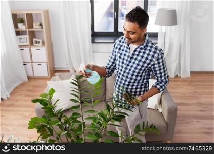 people, nature and plants care concept - smiling indian man spraying houseplant by water sprayer at home. man spraying houseplant with water at home