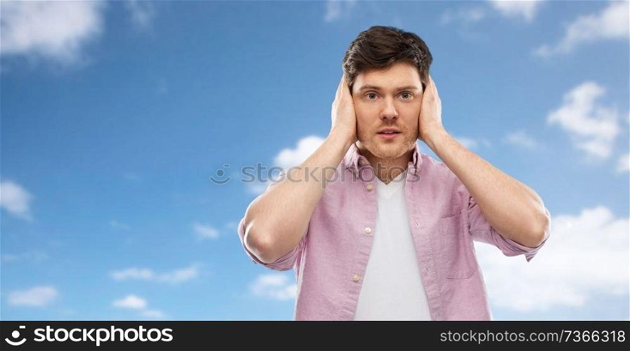 people, noise and stress concept - man closing ears by hands over blue sky and clouds background. man closing ears by hands over blue sky