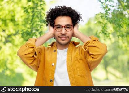 people, noise and stress concept - man in yellow jacket closing ears by hands over green natural background. man closing ears by hands on natural background