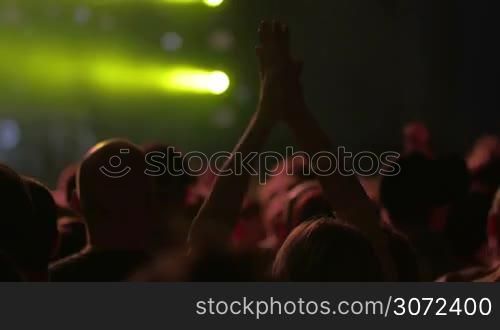 People on the concert. Focus on female hands applauding among the crowd. Digital screen and bright yellow spotlights in background, then the lights going down