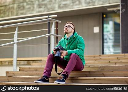 people, photography, technology, leisure and lifestyle - happy young hipster man holding digital camera with big lens and sitting on stairs in city