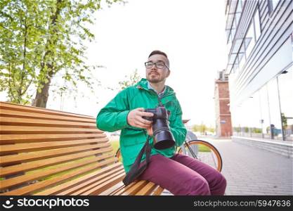 people, photography, technology, leisure and lifestyle - happy young hipster man holding digital camera with big lens and looking for subject on city street