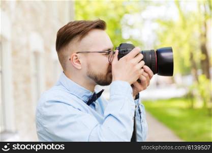 people, photography, technology, leisure and lifestyle - happy young hipster man holding digital camera with big lens taking picture on city street