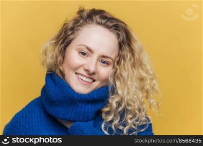People, positive emotions and beauty concept. Beautiful lovely young smiling female model in blue warm sweater, being satisfied with something, has pleasant smile on face, isolated on yellow wall