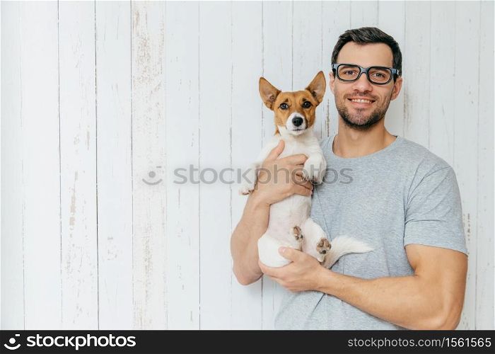 People, positiveness, animals and friendship concept. Happy unshaven male wears casual clothes, holds puppy, have walk together, stand together against white wooden wall with blank copy space