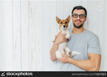 People, positiveness, animals and friendship concept. Happy unshaven male wears casual clothes, holds puppy, have walk together, stand together against white wooden wall with blank copy space