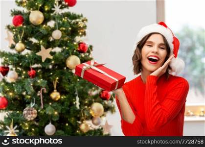 people, presents and holidays concept - happy smiling young woman in santa helper hat with red gift box over christmas tree on background. happy young woman in santa hat with red gift box