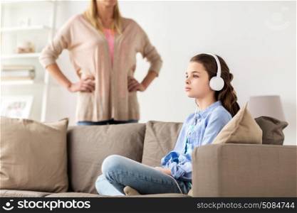 people, problem and family concept - girl with earphones sitting on sofa and angry mother at home. girl with earphones and angry mother at home. girl with earphones and angry mother at home