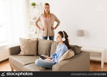 people, problem and family concept - girl with earphones sitting on sofa and angry mother at home. girl with earphones and angry mother at home