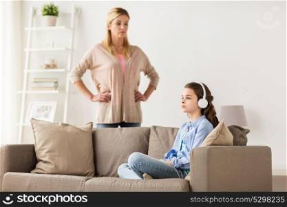 people, problem and family concept - girl with earphones sitting on sofa and angry mother at home. girl with earphones and angry mother at home