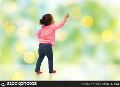 people, race, ethnicity and childhood concept - happy african american baby girl pointing finger to something from back over green summer holidays lights background
