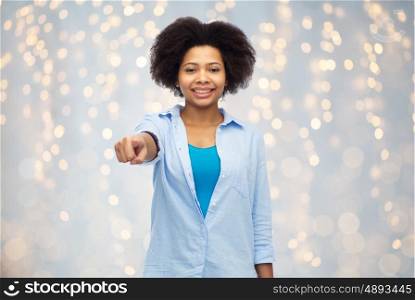 people, race, ethnicity, gesture and choice concept - happy african american young woman pointing finger to you over holidays lights background