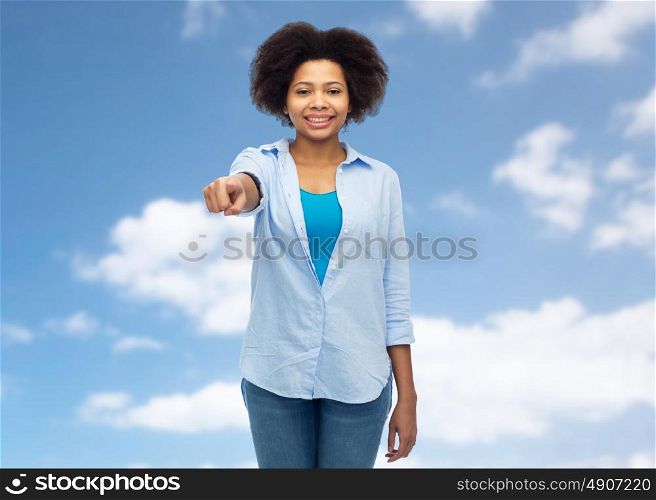 people, race, ethnicity, gesture and choice concept - happy african american young woman pointing finger to you over blue sky and clouds background. happy african woman pointing finger to you