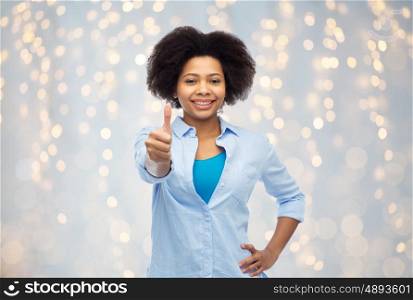 people, race, ethnicity, gesture and portrait concept - happy african american young woman showing thumbs up over holidays lights background
