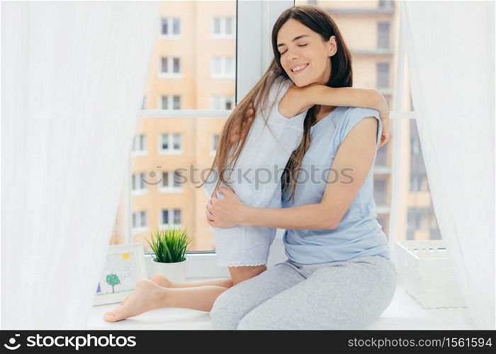 People, relationship and parenthood concept. Positive affectionate female with pleasant appearance, wears casual domestic clothes, embraces her small daughter, expresses love, upbrings her child