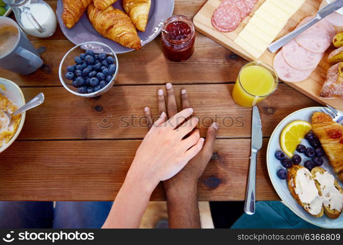 people, relationships and eating concept - multiracial couple holding hands while having breakfast at table full of food. couple hands on table full of food