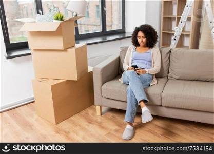 people, repair and real estate concept - sad african american woman with smartphone and boxes moving to new home. woman with smartphone and boxes moving to new home