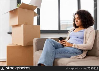 people, repair and real estate concept - sad african american woman with smartphone and boxes moving to new home. woman with smartphone and boxes moving to new home
