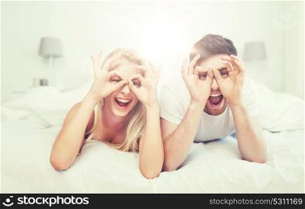 people, rest, love, relationships and fun concept - happy couple lying in bed at home and making finger glasses. happy couple lying in bed at home