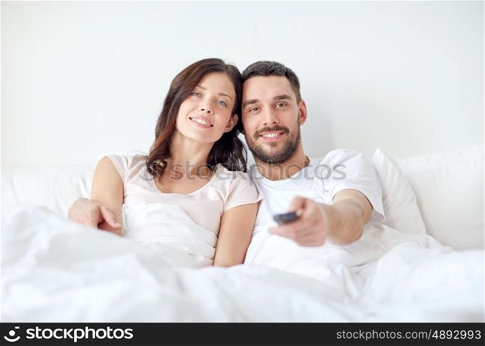 people, rest, love, relationships and television concept - happy couple with remote lying in bed at home and watching tv