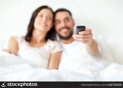 people, rest, love, relationships and television concept - happy couple with remote lying in bed at home and watching tv. happy couple lying in bed at home and watching tv