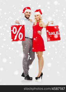 people, sale, christmas, winter and holidays concept - happy couple in santa hats with red shopping bags hugging over snow background