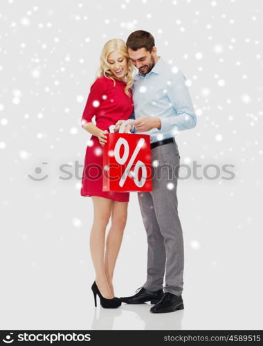 people, sale, christmas, winter and holidays concept - happy couple looking into red shopping bag with percentage sign over snow background