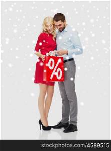 people, sale, christmas, winter and holidays concept - happy couple looking into red shopping bag with percentage sign over snow background