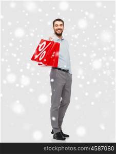 people, sale, christmas, winter and holidays concept - smiling man holding red shopping bag with percentage sign over snow background
