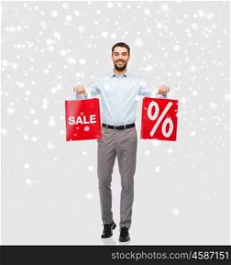 people, sale, christmas, winter and holidays concept - smiling man holding red shopping bags with percentage sign over snow background