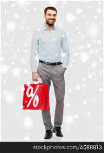 people, sale, christmas, winter and holidays concept - smiling man holding red shopping bag with percentage sign over snow background