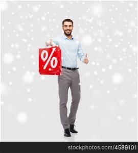 people, sale, christmas, winter and holidays concept - smiling man holding red shopping bag with percentage sign showing thumbs up over snow background