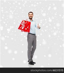 people, sale, christmas, winter and holidays concept - smiling man holding red shopping bags with percentage sign showing thumbs up over snow background