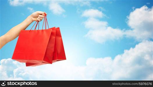 people, sale, consumerism, advertisement and commerce concept - close up of hand holding red blank shopping bags over blue sky and clouds background