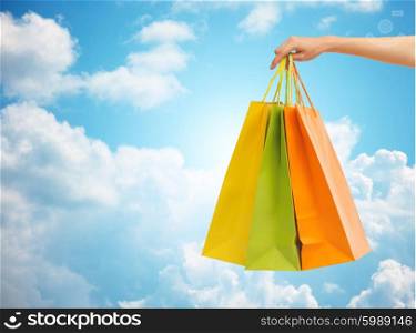 people, sale, consumerism and advertisement concept - close up of hand holding shopping bags over blue sky and clouds background