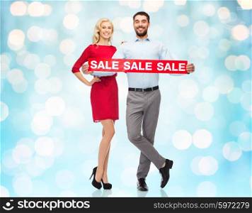 people, sale, discount and holidays concept - happy couple with red sale sign over blue holidays lights background