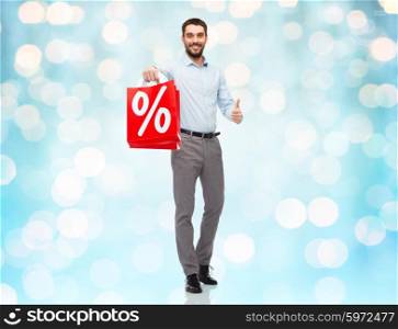 people, sale, discount and holidays concept - smiling man holding red shopping bags with percentage sign and showing thumbs up gesture over blue holidays lights background