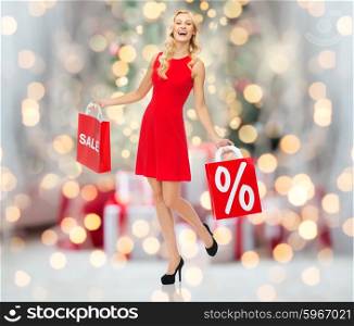 people, sale, discount and holidays concept - smiling woman in red dress with shopping bags over christmas tree lights background