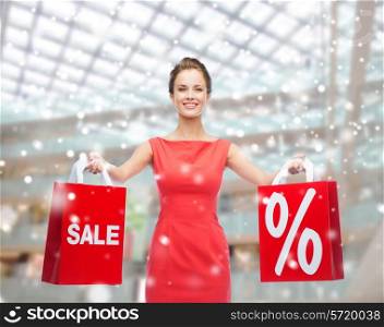 people, sale, gifts, christmas and holidays concept - smiling young woman in red dress holding bags with percent and sale sign over shopping center background
