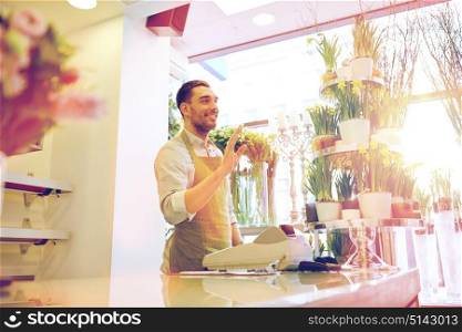 people, sale, retail, business and floristry concept - happy smiling florist man with cashbox standing at flower shop counter. florist man or seller at flower shop counter