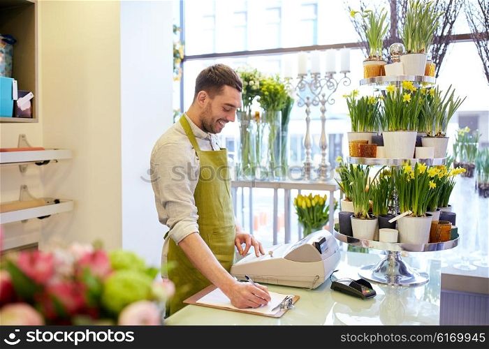 people, sale, retail, business and floristry concept - happy smiling florist man with clipboard and cashbox writing and making notes order at flower shop counter