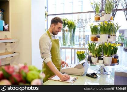 people, sale, retail, business and floristry concept - happy smiling florist man with clipboard and cashbox writing and making notes order at flower shop counter